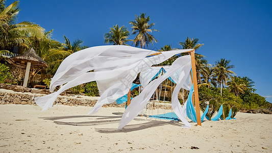 在靠近海洋的海滩上用鲜花装饰的婚礼拱门 她的白色和蓝色画布在风中飞舞 棕榈树叶子的装饰心 沙质热带海岸图片