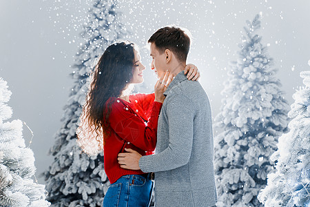 爱情故事在冬日庆祝新年的前夕 快乐的年轻夫妇在圣诞树附近拥抱和亲吻 笑脸男人和女人彼此相爱 飘落的雪花和亲吻 礼物 乐趣背景