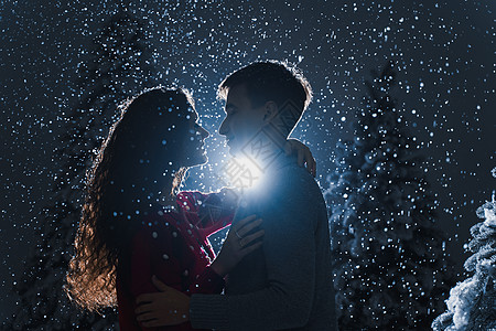 爱情故事落雪和深蓝色背景上落雪的亲吻 快乐的年轻夫妇在冬日庆祝新年的前夕在圣诞树附近拥抱和亲吻 农村 房子背景