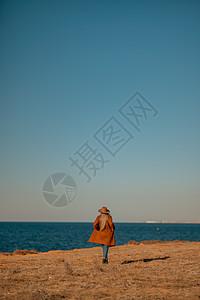 一个女人沿着海边的海岸散步 一位身穿棕色外套 戴着时尚妆容的帽子的优雅女士在海边散步 成人 天图片