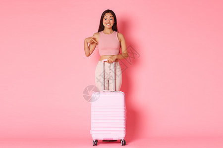 旅游 假期和假期的概念 穿着夏装的全身兴奋快乐的亚洲女孩手指向下 微笑着兴奋地站着 手提箱 粉红色背景图片