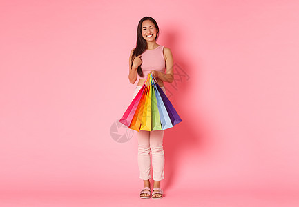 美丽而时髦的亚洲女孩购物狂的全长肖像 拿着购物袋指着自己 为她的假期买了夏装 站在粉红色背景图片