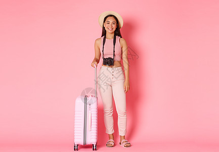 旅游 暑假 国外假期概念 身着帽子 微笑着的美丽女孩旅行者 带着相机和准备冒险的手提箱的游客 站在粉红色背景中图片