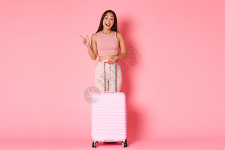 旅游 假期和假期的概念 身着夏装 探索国家 在国外享受夏天 向左指着 带着手提箱站着 粉红色背景的全长逗趣的美丽亚洲女孩图片