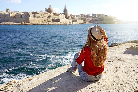 坐在海边石头上坐着帽子的美丽的年轻女士 看着马耳他瓦莱塔市令人惊叹的全景图片