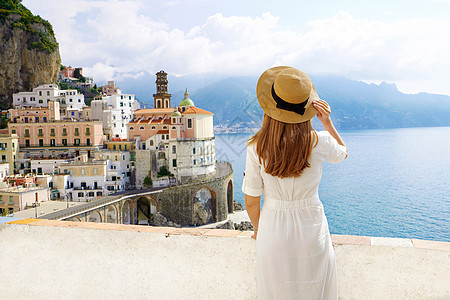 意大利旅游业 背景上与Atrani村一起戴草帽和白裙子的年轻女性背影 意大利阿马尔菲海岸图片