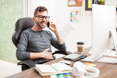 一个英俊的西班牙年轻人坐在家庭办公室时用智能手机说话的肖像校对图片