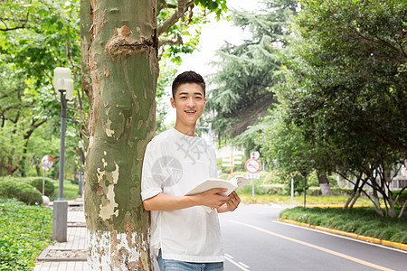 拿着书站在校园树下看书的男生图片