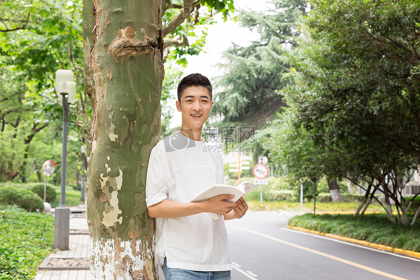 拿着书站在校园树下看书的男生图片