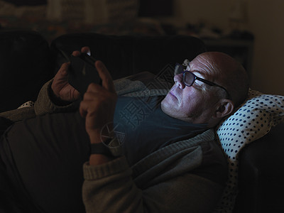 成熟的男人，晚上躺在沙发上，用智能手机，脸上有灯光图片