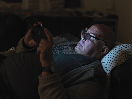 成熟的男人，晚上躺在沙发上，用智能手机，脸上有灯光图片
