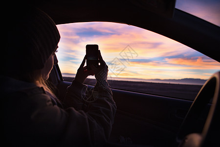 年轻女子在车内用智能手机拍摄日落图片