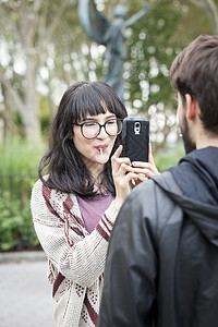 公园里用智能手机拍男朋友的年轻女子的肩上视图图片