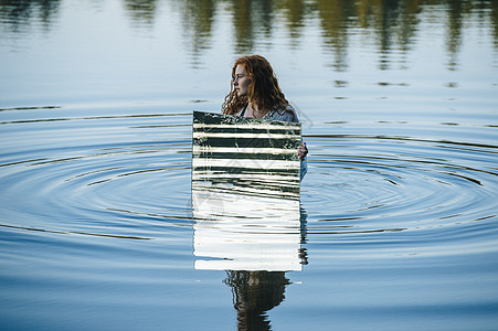 年轻女子站在湖中，举着镜子，水中的涟漪在镜子中反射图片