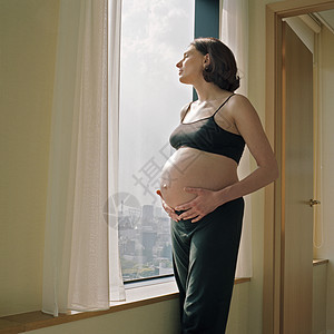 中年孕妇抱着肚子站在公寓窗户前的照片图片