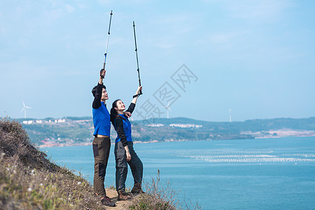 海边情侣海边山坡上举登山杖男女背景