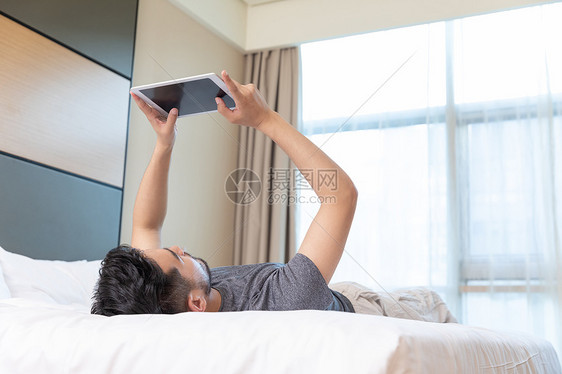 青年男性躺在酒店床上玩平板电脑图片