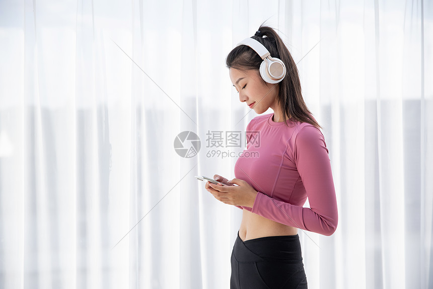 穿着运动装的年轻美女在窗边戴耳机听音乐图片