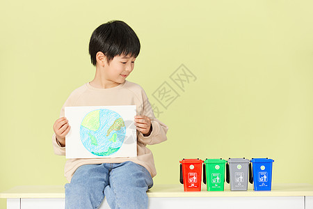 小男孩抱着手绘地球画看着垃圾分类桶背景图片