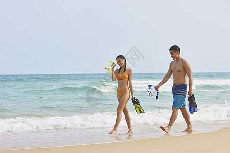 大海美女年轻情侣拿着潜水装备海边行走背景