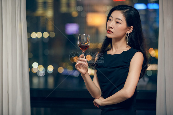 年轻美女夜晚端着红酒杯站在窗前图片