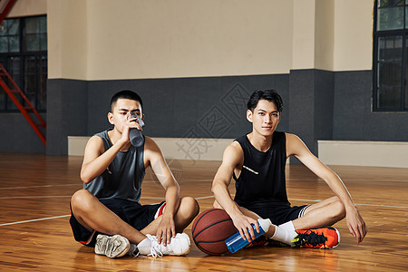 两个篮球运动青年坐在室内篮球场地板上喝水图片