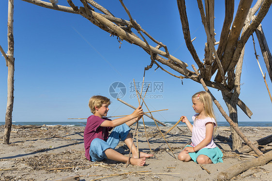 男孩和女孩在海滩上拿着浮木建造庇护所图片