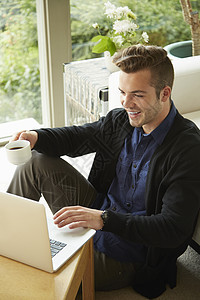坐在家中地板上喝咖啡看电脑微笑的男人肖像图片