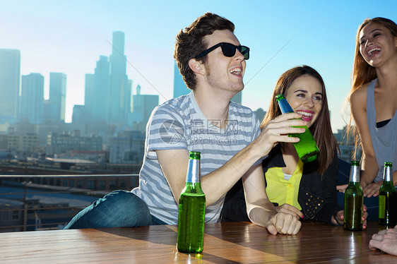 年轻男女饮啤酒在美国洛杉矶天际的屋顶酒吧大笑图片