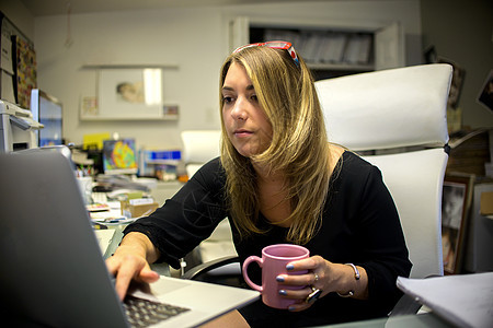 办公室中的年轻妇女坐在办公桌前用笔记本电脑拿咖啡杯图片