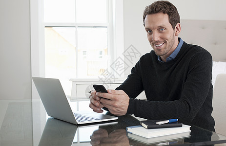 使用笔记本电脑和智能手机坐在桌边的中年成人男子图片