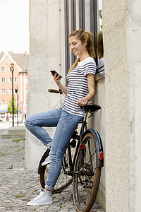 一边看年轻女靠着自行车一边仰着建筑外部一边看着智能手机微笑图片