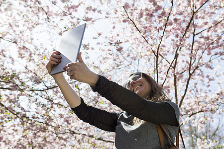 在樱花树下使用平板自拍的年轻女性图片