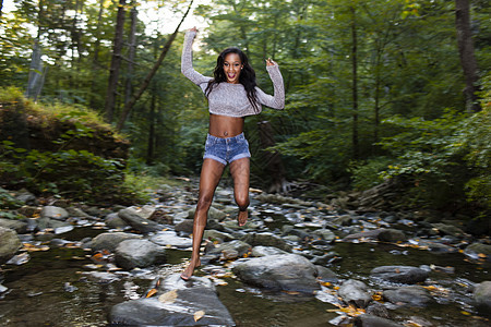 快乐的年轻女子跳过森林河流岩石的肖像图片