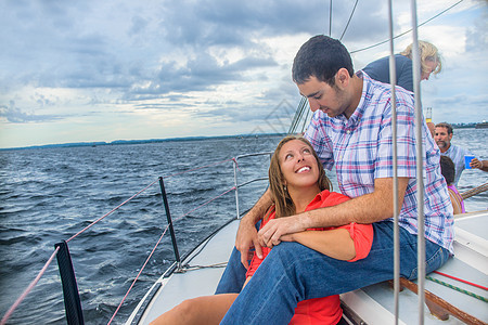 年轻女子坐在帆船上的年轻男子双腿中间回头看着彼此微笑图片