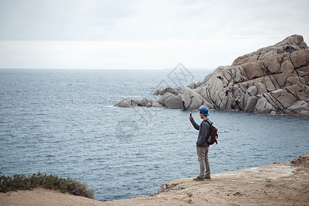 站在悬崖上的年轻人使用智能手机拍摄海洋照片图片