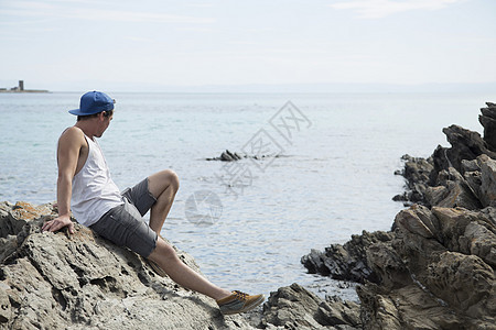 坐在岩石上看远视海洋的年轻男子全长侧面观意大利SintinoSardiniaSatdinia图片