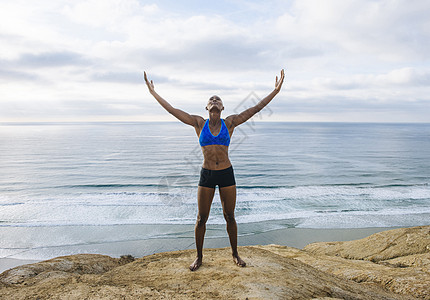 年轻女站在海边的岩石上举起手臂抬头看图片