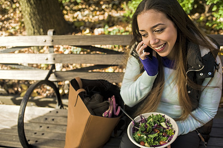 坐在公园长椅上的年轻女子在吃沙拉午餐时在智能手机上聊天图片