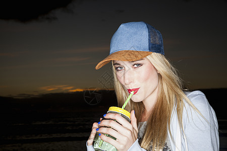 身戴棒球帽的年轻女子喝着饮料看着镜头微笑图片