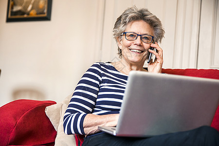 住在起居室沙发上的中年妇女使用笔记本电脑时智能手机说话图片
