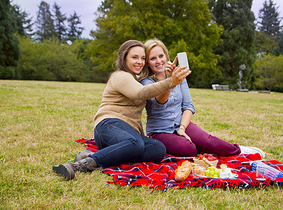 两个年轻女在公园享受野餐自拍使用智能手机图片