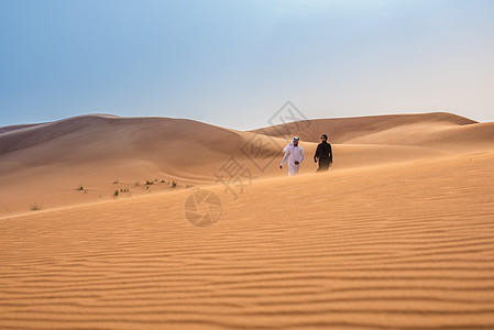 在阿拉伯联合酋长国迪拜沙漠丘上行走时穿着传统中东部服装的夫妇远景图片