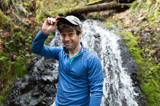在瀑布前面戴着棒球帽看镜头微笑的男人图片