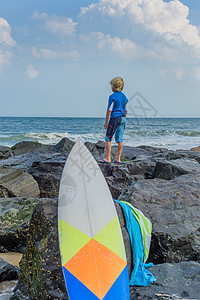 年轻男孩站在海边的岩石上看着风景表面的冲浪板图片