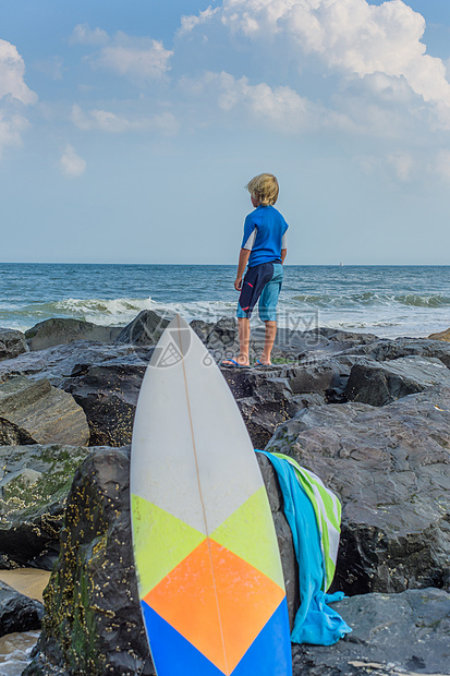 年轻男孩站在海边的岩石上看着风景表面的冲浪板图片