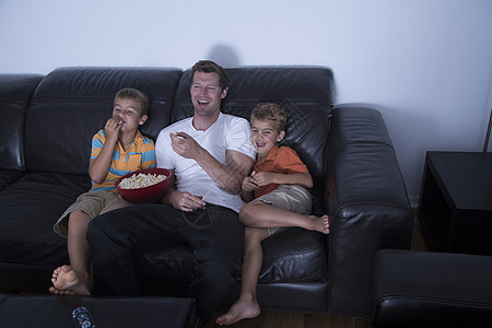 男人和两个儿子一边在沙发上吃爆米花一边笑着看电视图片