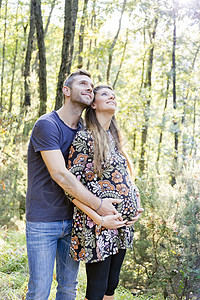 在森林拥抱的怀孕夫妇看微笑着抬头看图片