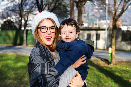 在公园里戴着圣诞帽的年轻女人抱着婴儿男孩图片