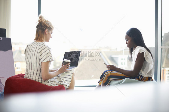 坐在豆袋上看笔记本电脑和智能手机的两名女商人图片